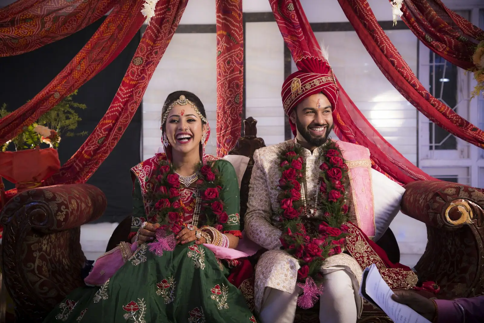 A couple seated together during their wedding ceremony, garlanded in red and gold